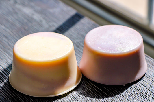 Two handcrafted round goat milk soaps from Milagro Goat Farm in natural sunlight on wood surface